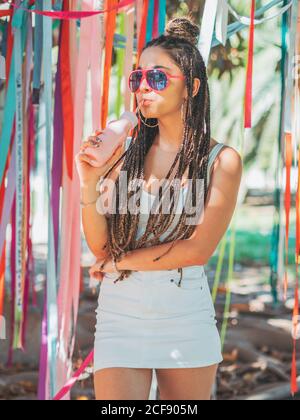 Stilvolle junge Frau trägt Sonnenbrille fürchten trinken Cocktail während draußen festival und wegschauen Stockfoto