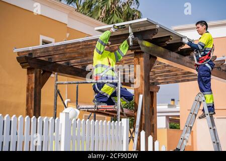 Gruppe von Arbeitern in Uniform und Helme Installation von Photovoltaik-Panels Auf dem Dach der Holzkonstruktion neben dem Haus Stockfoto