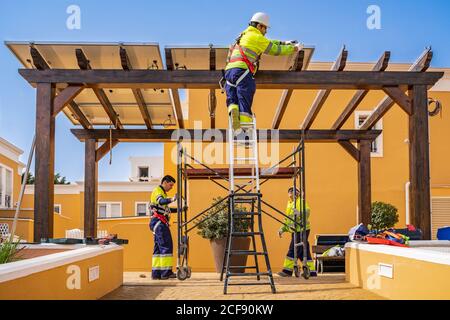 Gruppe von Arbeitern in Uniform und Helme Installation von Photovoltaik-Panels Auf dem Dach der Holzkonstruktion neben dem Haus Stockfoto