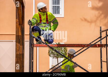 Professionelle Ingenieur in Uniform und Helm sitzt auf Gerüst und Wegschauen, während der Arbeit mit Kollegen auf die Wartung der elektrischen Ausstattung in der Nähe von Wohngebäuden Stockfoto