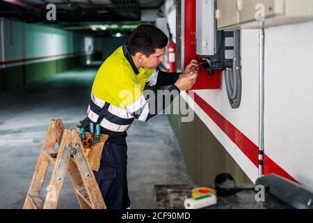 Seitenansicht des jungen professionellen männlichen Technikers mit Elektrowerkzeugen Reparatur und Überprüfung der Ausrüstung während der Arbeit im Gebäude Stockfoto