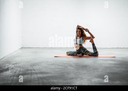 Seitenansicht der fitting female in Sportswear sitzend auf Matte Und Yoga in Eka Pada Rajakapotasana mit Mudra-Geste praktizieren Stockfoto