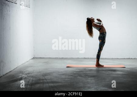 Seitenansicht einer ruhigen Frau mit Sport-Leggings und BH Balancing auf Bein im Stand backbend mit Gebet Hände und Suchen nach oben Stockfoto