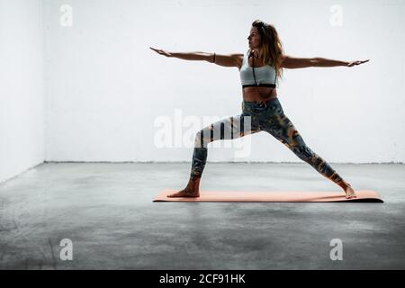 Seitenansicht einer ruhigen Frau mit Sport-Leggings und BH Stehend auf Matte Krieger zwei Pose wegschauen Stockfoto