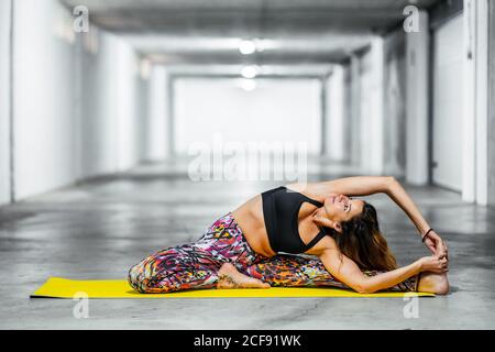 Seitenansicht einer erwachsenen Frau in drehter Kopf-Knie-Pose, während sie in der hellen Garagenhalle Yoga praktiziert und nach oben schaut Stockfoto