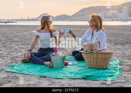 Fröhliche Mutter und Tochter lächeln und sehen sich an Während des Familientreffens auf einer Decke und klirrenden Tassen sitzend Picknick am Strand am Abend Stockfoto