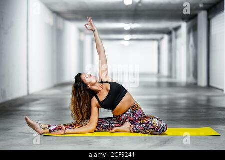 Seitenansicht einer erwachsenen Frau in drehter Kopf-Knie-Pose, während sie in der hellen Garagenhalle Yoga praktiziert und nach oben schaut Stockfoto