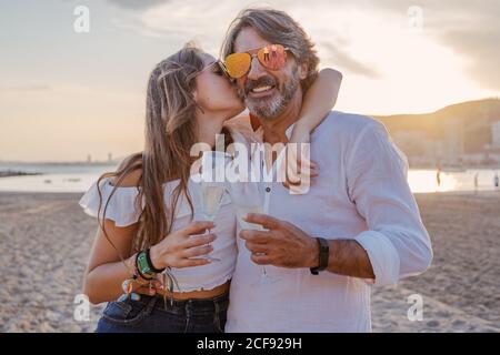 Reifer Mann umarmt und küsst junge Frau mit Wange, während er Toast vorschlägt und das Familientreffen am Sandstrand während der Dämmerung im Resort feiert Stockfoto