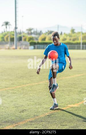 Ganzkörper schwarz Teenager in blau Sportswear Jonglieren roten Ball Auf dem Knie beim Training auf dem Fußballplatz Stockfoto