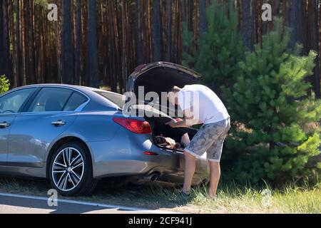 Der junge Erwachsene Mann lehnte sich über den Kofferraum eines Autos Geparkt entlang der Straße vor der Kulisse einer Kiefer Wald Stockfoto