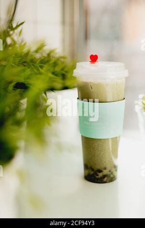 Plastikbecher mit Herz von leckeren heißen Matcha-Tee auf Tisch in der Nähe von Topfpflanzen im asiatischen Restaurant Stockfoto