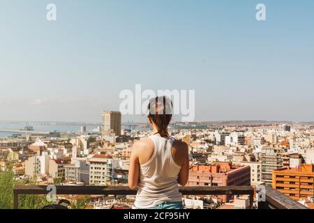 Rückansicht einer jungen Frau im weißen T-Shirt, die an einem hellen Tag in Alicante, Spanien, die Aussicht auf die Stadt vom Balkon genießt Stockfoto