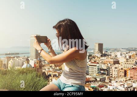 Seitenansicht einer jungen Frau im weißen T-Shirt, die an einem hellen Tag in Alicante, Spanien, Stadtansichten vom Balkon fotografiert Stockfoto