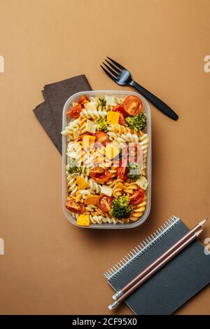 Draufsicht auf Kunststoff-Lunchbox mit Pasta, Tomaten, Käse und Brokkoli platziert durch schwarze Kunststoffgabel und Notizblock mit Bleistiften auf braunem Hintergrund Stockfoto