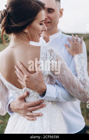 Braut in weißem Kleid steht mit dem Rücken, ihr Bräutigam umarmt sie vor der Kulisse der Berge. Stilvolle Braut und schöne Braut stehen auf der Klippe. Stockfoto