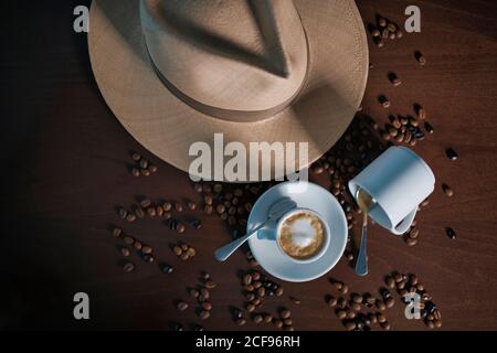 Von oben köstliches duftendes braunes Getränk mit weißem Schaum in Keramikbecher mit Teelöffel auf runden Untertasse unter geröstetem Kaffee Bohnen neben Hut mit Krempe und leerer Tasse auf Holztisch Stockfoto