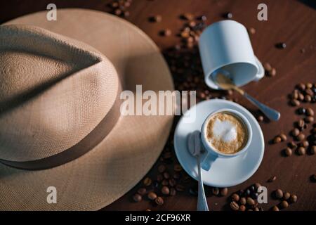 Von oben köstliches duftendes braunes Getränk mit weißem Schaum in Keramikbecher mit Teelöffel auf runden Untertasse unter geröstetem Kaffee Bohnen neben Hut mit Krempe und leerer Tasse auf Holztisch Stockfoto