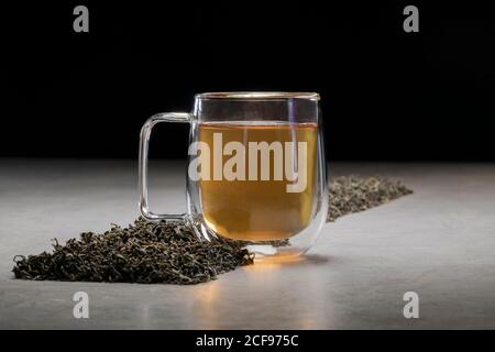 Aromatisches Getränk in Glas Becher mit Haufen von getrockneten angeordnet Teeblätter auf dem Tisch auf schwarzem Hintergrund Stockfoto