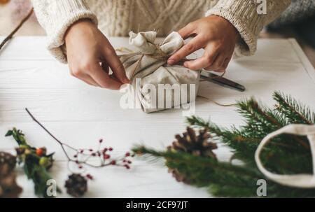 Weibliche Hände wickeln stilvolles weihnachtsgeschenk in Leinenstoff auf weißen rustikalen Tisch mit grünen Zweig, Kegel, Schere und Garn. Frau im gemütlichen sw Stockfoto