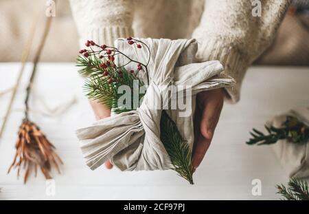 Weihnachtsgeschenk ohne Verschwendung. Hände halten stilvolle Geschenk in Leinenstoff mit grünen Tannenzweig und roten Beeren auf rustikalem Hintergrund eingewickelt. Kunststoff frei Stockfoto