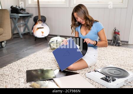Glücklich junge weibliche Meloman in lässigen Outfit sitzt auf Teppich Auf dem Boden neben der Gitarre und Musik mit Vinyl hören Disc- und Plattenspieler, während Sie sich zu Hause ausruhen Stockfoto