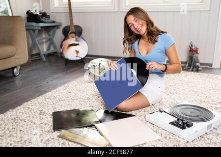 Glücklich junge weibliche Meloman in lässigen Outfit sitzt auf Teppich Auf dem Boden neben der Gitarre und Musik mit Vinyl hören Disc- und Plattenspieler, während Sie sich zu Hause ausruhen Stockfoto