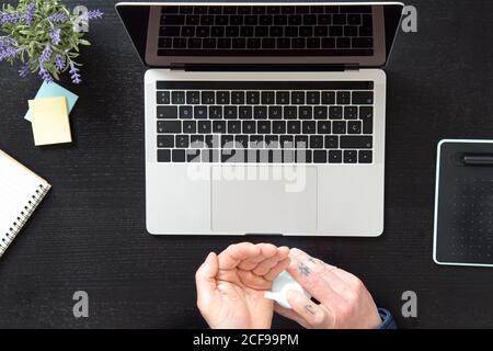 Von oben Ernte männlichen Behandlung Hände mit Desinfektionsmittel zum Schutz Von Coronavirus während der Arbeit auf Laptop zu Hause während der Quarantäne Stockfoto