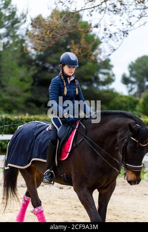 Teen Mädchen Jockey im Helm Reiten braun Pferd unter Baum Zweige auf Dressurviereck während der Ausbildung in der Reitschule Stockfoto