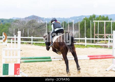 Rückansicht von unkenntlich teen Mädchen im Helm sitzt auf Pferd zurück und springen über Hürde während des Trainings auf Dressur arena der Reitschule Stockfoto