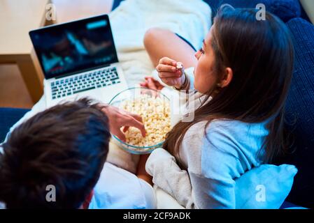 Fröhliche junge Männer und Frauen in legerer Kleidung essen Popcorn und schauen sich Filme auf dem Laptop an, während sie sich zu Hause auf dem gemütlichen Sofa ausruhen Stockfoto