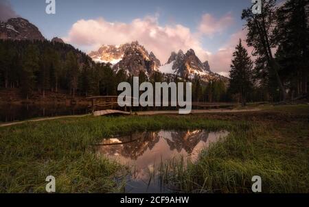 Befriedende Landschaft des dichten Waldes, entlang dem Weg mit Kleine Brücke neben runden Teich breitet sich auf üppige Grüner Rasen spiegelt Himmel und Berge in den Dolomiten Itlay Stockfoto