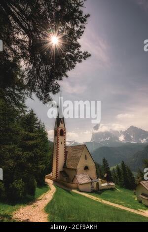 Waldwege, die zu einer gemütlichen kleinen Kirche auf einem grünen Hügel führen Unter heller Sonne leuchtet Licht durch dunkles Laub wolkenverhangener Himmel in den Dolomiten Italien Stockfoto