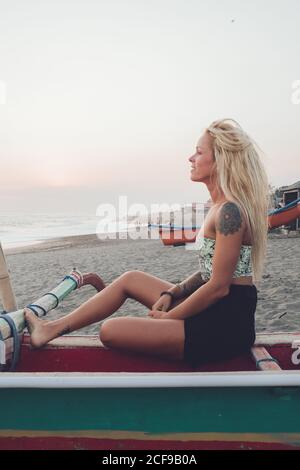 Seitenansicht der positiven jungen Dame im lässigen Kleid sitzen Auf einer Holzbank am Sandstrand und mit Blick auf das wehende Meer Im Sommer bei Sonnenuntergang Stockfoto