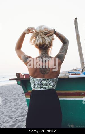 Rückansicht einer unkenntlich schlanken jungen Frau in Kleid und mit Körper in Tattoos, die mit erhobenen Händen am Kopf am tropischen Strand steht und während des Sonnenuntergangs das winkende Meer bewundert Stockfoto