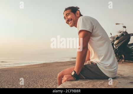 Von unten Seitenansicht des positiven aktiven ethnischen Kerl mit Motorrad auf Steinzaun sitzen und den Sonnenuntergang auf leer genießen Sandstrand Stockfoto