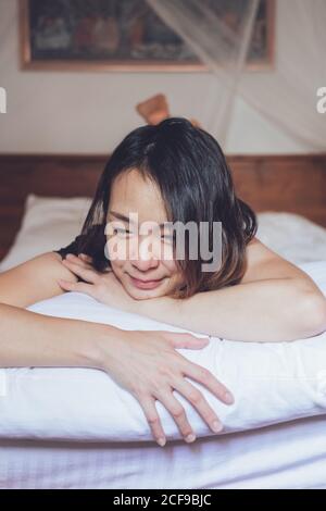 Eine optimistische asiatische Frau schaut weg, liegt auf einem bequemen Bett und lacht, während sie morgens zu Hause auf dem Bett liegt Stockfoto