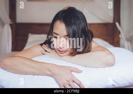 Eine optimistische, nachdenkliche asiatische Frau, die morgens zu Hause auf einem bequemen Bett liegt Stockfoto