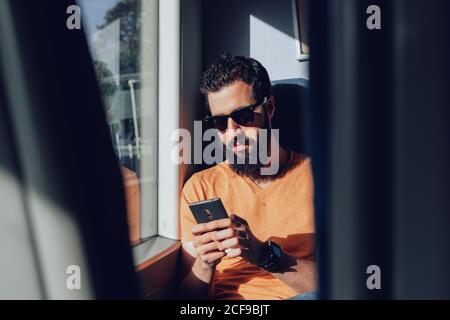 Ruhiger bärtiger Mann in legerer Kleidung und Sonnenbrille mit Handy Telefon, während auf Beifahrersitz in der Nähe Fenster in modernen sitzen Trainieren Stockfoto