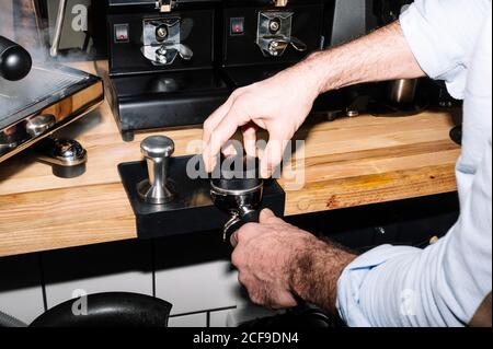 Großer Winkel des männlichen Barista, der gemahlenen Kaffee in einem Portafilter presst Bei der Zubereitung von frischen Getränken in einer professionellen Kaffeemaschine Stockfoto