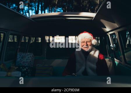 Lächelnder älterer Mann im Kostüm des Weihnachtsmannes, der innen sitzt Ein alter Lieferwagen in der Nähe verpackt Geschenke an sonnigen Tag suchen Bei der Kamera Stockfoto