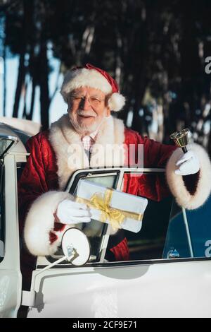 Lächelnder älterer Mann im Kostüm des Weihnachtsmannes, der innen steht Eine alte van Tür mit eingewickelten Geschenk und Glocke auf Sonniger Tag mit Blick auf die Kamera Stockfoto
