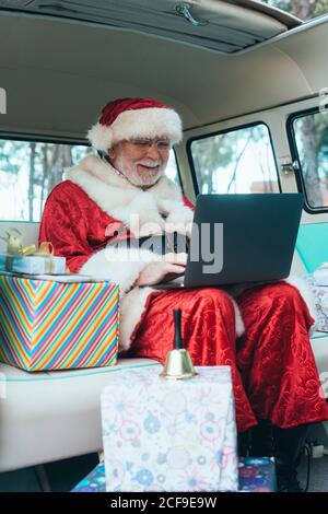 Fröhlicher Mann im Kostüm des Weihnachtsmannes, der im Van sitzt Mit Laptop und bunten Geschenken an sonnigen Tagen Stockfoto