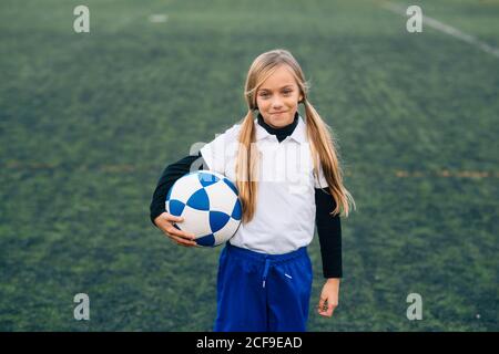 Fröhliche preteen Mädchen in weiß und blau Uniform mit Fußball Ball lächelt die Kamera an, während er allein auf dem grünen Feld steht Im modernen Sportverein Stockfoto