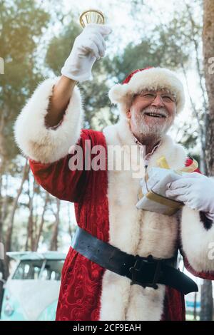 Fröhlicher älterer Mann im Kostüm des Weihnachtsmannes stehend mit Geschenk und Glocke in Handschuhen, die auf die Natur blicken Hintergrund Stockfoto