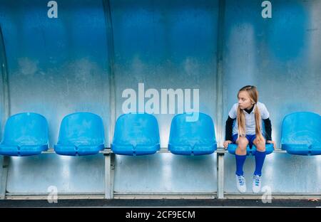 In voller Länge frustriert preteen Mädchen in Fußball-Uniform allein sitzen Auf blauem Kunststoffsitz nach Spielausfall im Sportverein Stockfoto