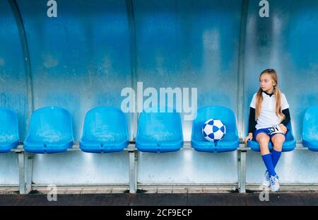 In voller Länge frustriert preteen Mädchen in Fußball-Uniform allein sitzen Auf blauem Kunststoffsitz nach Spielausfall im Sportverein Stockfoto