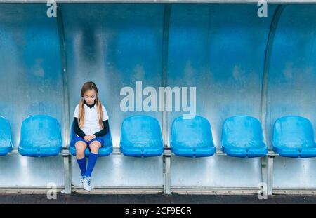 In voller Länge frustriert preteen Mädchen in Fußball-Uniform allein sitzen Auf blauem Kunststoffsitz nach Spielausfall im Sportverein Stockfoto