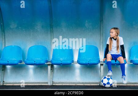 In voller Länge frustriert preteen Mädchen in Fußball-Uniform allein sitzen Auf blauem Kunststoffsitz nach Spielausfall im Sportverein Stockfoto