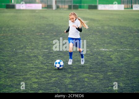 Teenager Mädchen in weiß und blau Uniform und Stollen laufen Und die Vorbereitung auf den Ball zu treten, während Fußball allein auf Green Field im modernen Sportclub Stockfoto