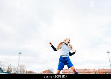 Aufgeregt Teenager-Mädchen in weiß und blau Uniform springen mit Arm angehoben und schreien, während der Sieg im Fußballspiel zu feiern Auf dem grünen Feld im modernen Sportverein Stockfoto